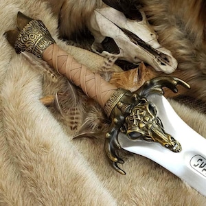 May include: A decorative sword with a tan leather-wrapped handle and ornate gold-colored metal accents. The blade is white with a small inscription. The sword features a detailed gold-colored metal hilt with a skull design. The background includes fur and a skull.