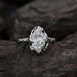 Puede incluir: Un anillo de plata con una piedra de cristal blanco translúcido, cruda y sin tallar, sujeta en un engaste de garras. La banda metálica tiene una apariencia texturizada y retorcida, con un diseño orgánico de tonos plateados metálicos.