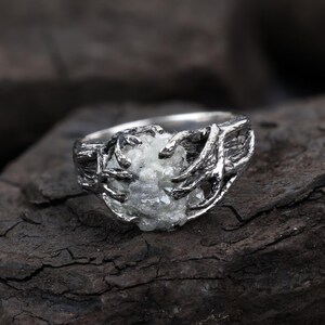 May include: A silver ring featuring a textured, dark metal band designed to resemble gnarled tree branches or roots. The band holds a central, rough-cut, translucent white gemstone that appears jagged and irregular in its natural, unpolished state.