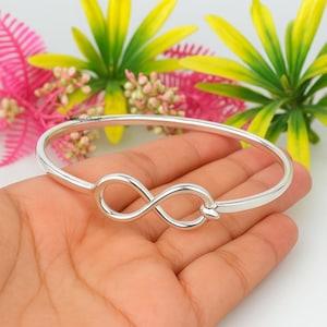 Può includere: Un braccialetto rigido color argento con un simbolo dell'infinito al centro. Il braccialetto ha una finitura liscia e lucida, con un design semplice ed elegante. Lo sfondo presenta fiori colorati.