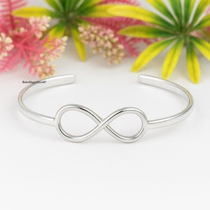 May include: A silver-colored cuff bracelet featuring an infinity symbol at the center. The open-ended design allows for easy wear. The bracelet is displayed against a backdrop of colorful flowers and foliage.