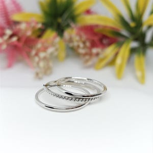 May include: Three silver rings, one plain, one with a twisted rope design, and one smooth. The rings are stacked on a white surface, with blurred flowers in the background. A simple jewellery set.
