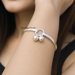 May include: A silver-toned bracelet with a circular clasp and two small, round charms. The bracelet is worn on a person's arm. The text "BohoMagicSilver07" is visible on the arm. The jewelry is simple and elegant.