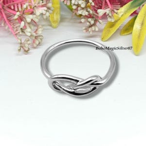 May include: A silver-colored ring with a knot design. The ring is made of a shiny metal and features a detailed knot in the center. The ring is displayed on a white surface with colorful flowers in the background. The text "BohoMagicSilver07" is visible.