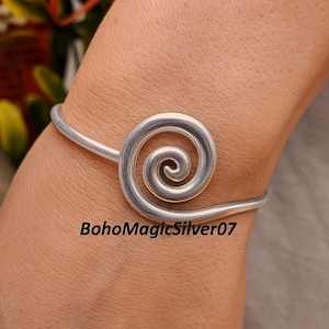May include: A silver-toned cuff bracelet featuring a spiral design. The bracelet has a simple, elegant style. The text "BohoMagicSilver07" is printed on the image.