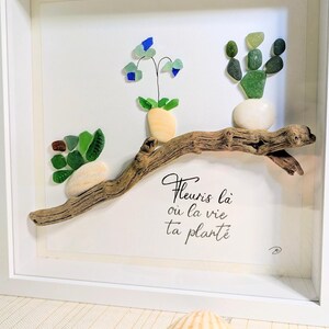 Peut inclure: Une œuvre d'art murale encadrée représentant trois plantes composées de galets et de verre poli fixées sur un morceau de bois flotté. Le texte "Fleuris là où la vie ta planté" est écrit en noir. Deux coquillages sont posés au premier plan.