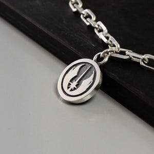 May include: Silver chain necklace with a pendant. The pendant is oval-shaped and features a detailed emblem with a star and wings design. The chain and pendant are displayed on a dark surface.