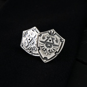 Puede incluir: Dos gemelos plateados en forma de escudo con intrincados diseños grabados. Los gemelos se colocan sobre un fondo de tela negra, creando un contraste que resalta los detalles del trabajo en metal. El diseño presenta un escudo estilizado.
