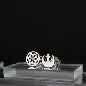 May include: A pair of silver-colored stud earrings. One earring features a circular design with a symmetrical pattern, while the other has a crescent shape with a stylized symbol. The earrings are displayed on a clear, rectangular surface against a black background.