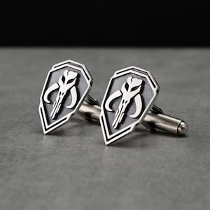Puede incluir: Un par de gemelos plateados con un diseño en forma de escudo y un emblema de calavera blanco. Los gemelos tienen un acabado pulido y un estilo clásico, adecuados para vestimenta formal.