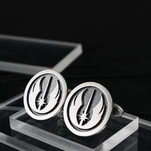 Könnte beinhalten: Silberne Manschettenknöpfe mit ovalem Design, die ein stilisiertes Emblem aus Lichtschwert und Flügeln zeigen. Die Manschettenknöpfe werden auf transparenten Acrylblöcken vor dunklem Hintergrund präsentiert.