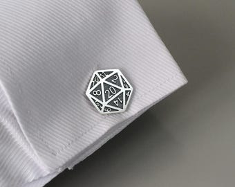 May include: A silver-colored cufflink in the shape of a twenty-sided die, with numbers etched in black. The cufflink is attached to a white dress shirt cuff, with the number 20 prominently displayed. The overall theme is gaming.