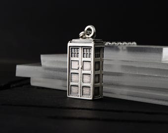 May include: A silver-toned metal pendant shaped like a rectangular police box with a flat roof, a small top light, and a grid-patterned door design. The pendant features a jump ring at the top for attachment to a chain, with a weathered finish.