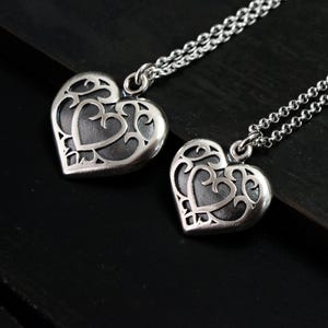 May include: Two silver heart-shaped pendant necklaces. Each pendant features a filigree design around a solid heart shape. The necklaces are on a silver chain. The background is a dark, textured surface.