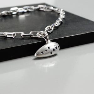 Puede incluir: Un collar plateado con un diseño de cadena. El collar presenta un colgante en forma de óvalo alargado con múltiples agujeros oscuros. El collar se exhibe sobre una superficie rectangular oscura.