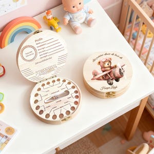 May include: Two wooden baby tooth keepsake boxes. One is open, showing tooth-shaped compartments and a space for a baby tooth. The other box has a teddy bear in a biplane and the name "Harper Leigh."
