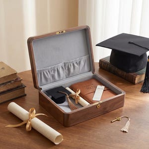 May include: A wooden graduation keepsake box with a gray interior, containing a miniature graduation cap, diploma, and a leather-bound journal with "CLASS OF 2026" embossed on it. A full-sized graduation cap and a rolled diploma are also visible.