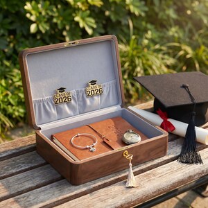 Könnte beinhalten: Geöffnete Holz-Andenkiste mit grauem Innenraum, gefüllt mit einem braunen Lederjournal, einem silbernen Armband und einer Taschenuhr. Zwei goldene Anstecknadeln mit Abschlusskappen und dem Text "CLASS OF 2026" befinden sich im Inneren. Eine Abschlusskappe und ein Diplom liegen daneben.