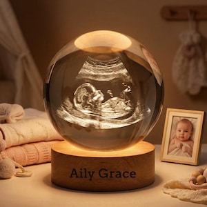 Op de afbeelding: Een kristallen bol lamp met een echo-afbeelding van een baby, rustend op een houten voet met de inscriptie "Aily Grace". Een ingelijste babyfoto staat naast de lamp. De lamp geeft een warm licht.