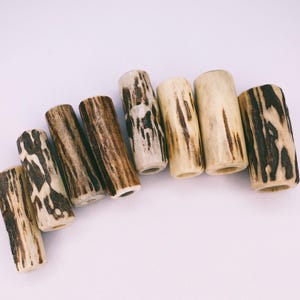 Op de afbeelding: Een verzameling van zeven cilindrische geweikralen. De kralen hebben een natuurlijke, crèmekleurige basis met donkerbruine, organische patronen. Elke kraal is in het midden doorboord, wat de natuurlijke textuur en kleurvariaties laat zien.