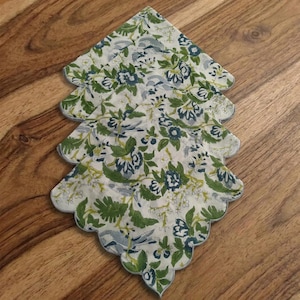May include: Four scalloped-edge cloth napkins with a floral pattern. The napkins feature a white background with green, blue, and gray floral designs. The napkins are stacked on a wooden surface.