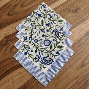 Puede incluir: Una pila de cuatro servilletas de tela cuadradas con una base blanca y un patrón de enredadera floral en azul y verde. Cada servilleta tiene un borde en zigzag de chevrón azul y blanco a lo largo de los lados, creando un efecto visual superpuesto.