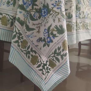 May include: White tablecloth with a floral pattern in blue, green, and olive. The border features a striped design in light blue and white. The tablecloth is draped over a wooden table.
