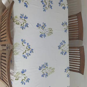 May include: A white rectangular tablecloth featuring a repeating pattern of blue flowers with green stems and leaves, draped over a wooden dining table surrounded by four wooden chairs with slatted backrests, viewed from an overhead perspective.
