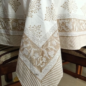 May include: A white tablecloth featuring a tan block-printed pattern of stylized trees and floral motifs. The border displays a detailed vine design with flowers, complemented by a striped edge pattern in a matching tan and white color scheme.