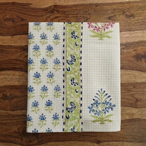 May include: A white waffle-weave kitchen towel featuring a repeating pattern of small blue flowers with green stems, a central vertical band with intricate green and blue floral motifs, and a single large blue floral spray on the right side.