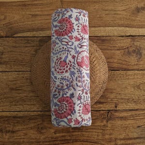 May include: A cylindrical roll of white fabric featuring a repeating floral pattern in shades of pink and light purple. The textile displays stylized blossoms and leafy vines arranged in a vertical orientation across the surface of the material.