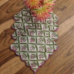 Puede incluir: Un conjunto de tres manteles individuales decorativos con bordes festoneados. Cada uno presenta un fondo blanco con un patrón floral verde y rosa. Los bordes están ribeteados con un borde rosa. Flores artificiales rosas y amarillas encima.