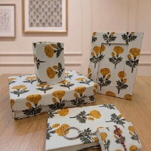 May include: A set of desk accessories featuring a white fabric covering with a repeating yellow marigold and green leaf pattern. The collection includes a rectangular box, a cylindrical pen holder, a picture frame, a notebook, and a small square box.
