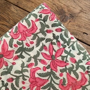 May include: A folded piece of off-white fabric featuring a repeating floral pattern. The design consists of large, vibrant pink lily-like flowers with dark green stems and leaves, arranged in a continuous botanical print across the textile surface.