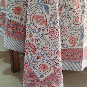 May include: A white tablecloth with a floral pattern in shades of pink and blue. The border features a pink floral design with a blue and white striped edge. The tablecloth is draped over a wooden table.