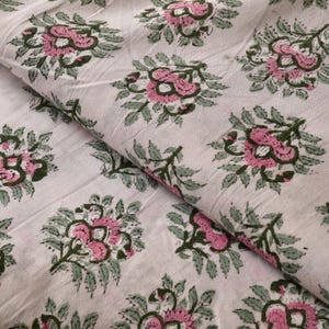 Puede incluir: Tela blanca con un estampado floral repetitivo. El diseño presenta flores rosas con hojas y tallos verdes. La tela está doblada, mostrando la textura y el patrón del material. El estampado floral está espaciado uniformemente en la tela.