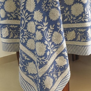 May include: A blue tablecloth with a white floral pattern and a striped border. The floral design features large, stylized flowers and leaves. The tablecloth is draped over a wooden table.