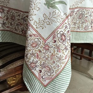 May include: A white tablecloth featuring a block-printed floral pattern in shades of pink, brown, and green. The fabric includes a decorative border with a repeating striped design in light green and white, framing the central botanical motif.