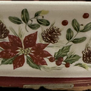 Puede incluir: Una fuente rectangular de cerámica blanquecina con un diseño festivo de invierno. La fuente presenta una flor de pascua, piñas, acebo y bayas en rojo, verde y marrón. La fuente se apoya sobre una base estampada en rojo y blanco.