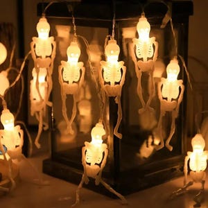 May include: String of Halloween skeleton lights. Each white skeleton has a glowing light bulb head. The skeletons hang in front of a black lantern. The lights are a festive decoration for Halloween.