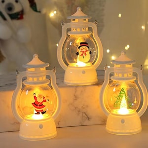 Puede incluir: Tres faroles navideños blancos con globos transparentes, cada uno con un personaje festivo diferente: un muñeco de nieve, Papá Noel y un árbol de Navidad con el texto "Merry Christmas". Los faroles emiten una luz cálida y brillante.