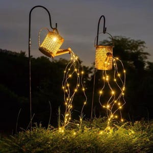 May include: Two decorative outdoor solar lights shaped like watering cans. The lights are gold-colored with a perforated design, and the "water" is represented by strings of small, warm-toned LED lights. They are mounted on black metal stakes.
