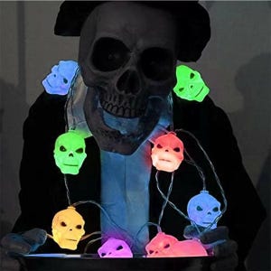 May include: A Halloween decoration featuring a large skull with a black hat, decorated with a string of glowing skull-shaped lights. The lights are in various colours, including green, blue, orange, and yellow, creating a spooky atmosphere.