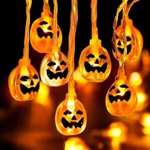 Könnte beinhalten: Eine Lichterkette mit orangefarbenen Kürbislichtern mit geschnitzten Gesichtern, ideal für Halloween-Dekorationen. Die beleuchteten Lichter erzeugen ein warmes Leuchten vor einem dunklen Hintergrund. Die Kürbisse hängen an einem dünnen, orangefarbenen Draht.