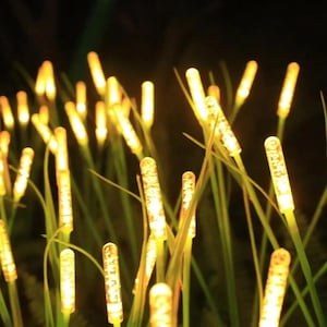 May include: Close-up of illuminated outdoor decorative lights. The lights are cylindrical with a textured surface, emitting a warm yellow glow. They are arranged among green blades of grass, creating a natural, inviting ambiance.