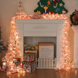 Peut inclure: Un manteau de cheminée blanc décoré pour Noël avec une guirlande de fleurs roses et de lumières. Trois boîtes cadeaux sur le thème de Noël sont empilées devant la cheminée. Un sac en papier avec le texte "I MISS DAI" est visible.