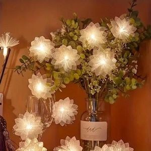 May include: A display of illuminated, white, flower-shaped lights. The lights are arranged around a vase of greenery and in a wine glass. The lights have a soft, glowing effect, creating a warm and inviting ambiance.