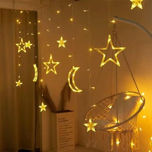 May include: Warm yellow star and crescent moon string lights hang from the ceiling. A macrame swing chair is lit by the lights. The lights create a cozy, inviting atmosphere. The lights are perfect for a bedroom or living room.
