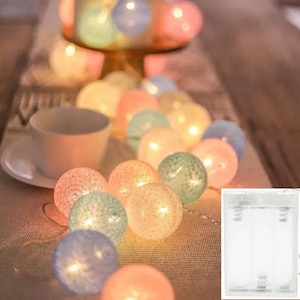 Può includere: Una stringa di luci decorative con palline di cotone multicolore, tra cui rosa, blu, verde e giallo. Le luci sono infilate su un filo, creando un bagliore caldo. Sullo sfondo si vedono una tazza e un piattino bianchi.