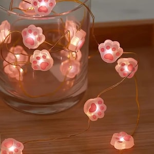 Puede incluir: Una cadena de luces decorativa con luces rosas en forma de huellas de pata. Las luces están dispuestas en un tarro de cristal transparente y sobre una superficie de madera. Cada huella tiene un centro rosa y emite un brillo cálido.
